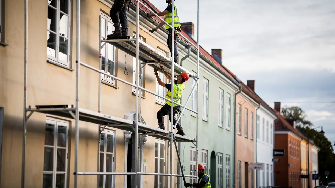 Stilladsmontør i sikkerhedssele monterer dæk på et facadestillads foran et gult murstenshus